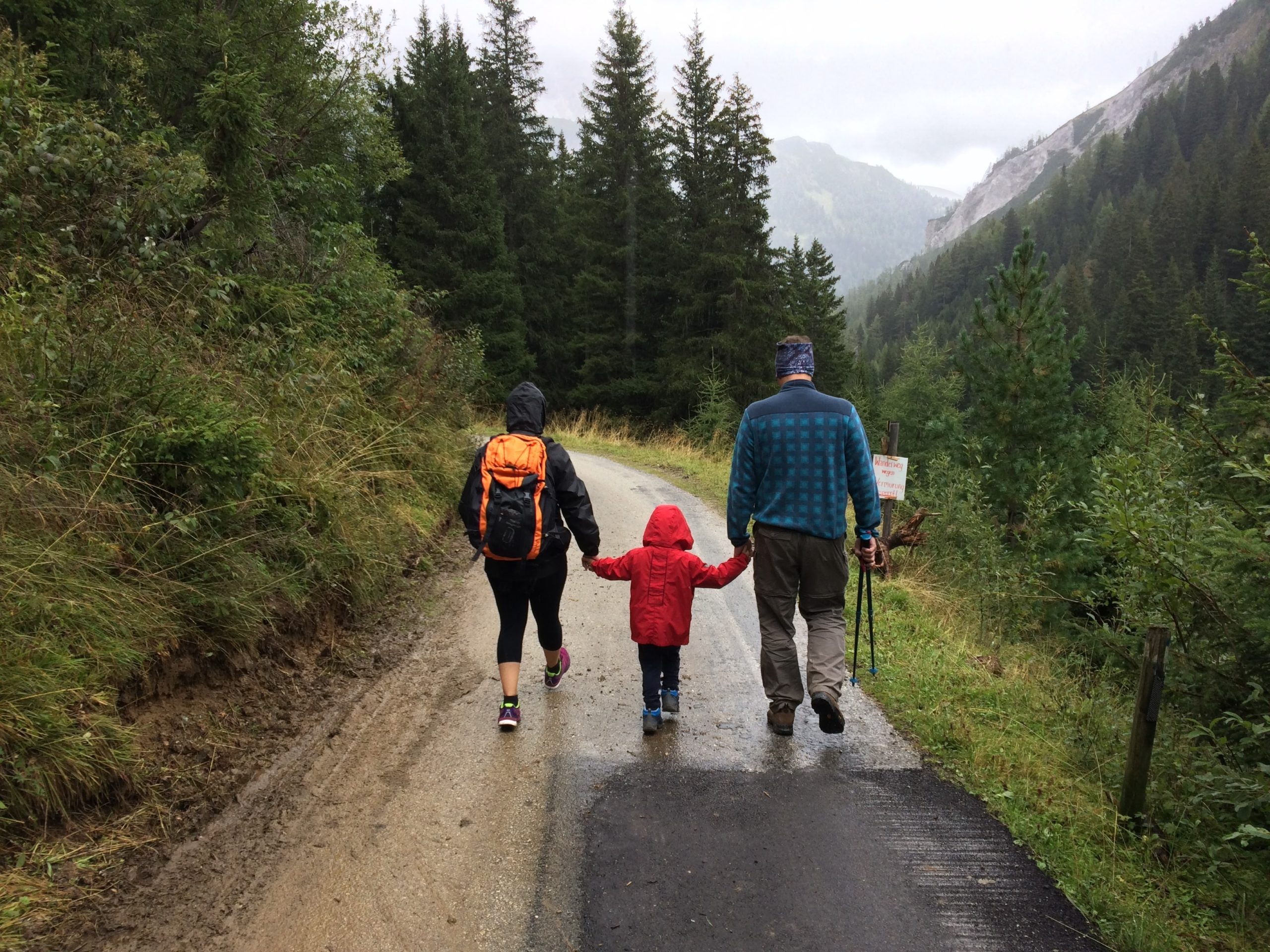 Two parents walking in the rain and with child inbetween them. Child has bright red raincoat on and everyone is holding hands. Walking along mountainous road