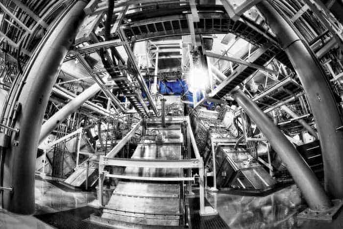 Photo of the target chamber at the National Ignition Facility. The photo is mostly in black-and-white, with the chamber showing up as a bright blue sphere at the centre of the photo. The chamber is surrounded by laser beamlines, which are long metal boxes that enclose the laser beam and optics. The photo also shows metal access stairs and catwalks, and metal structures supporting the beamlines and instrumentation