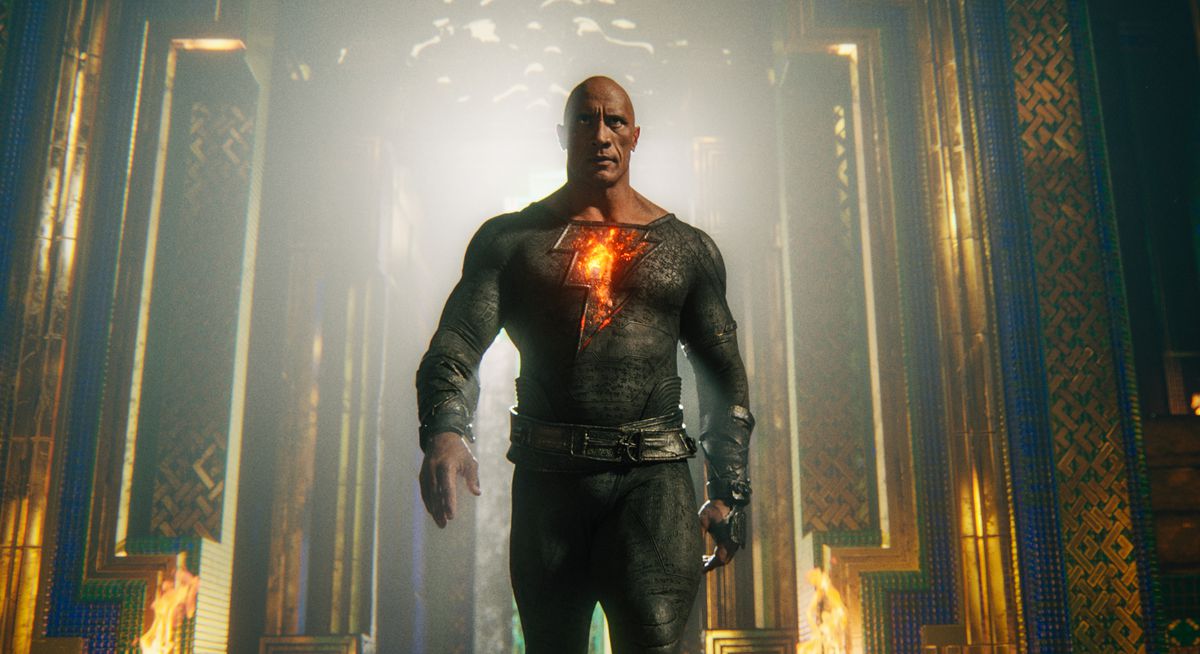 Dwayne Johnson strides toward the camera in a form-fitting black suit with a glowing lightning bolt on the chest as the title character in Black Adam