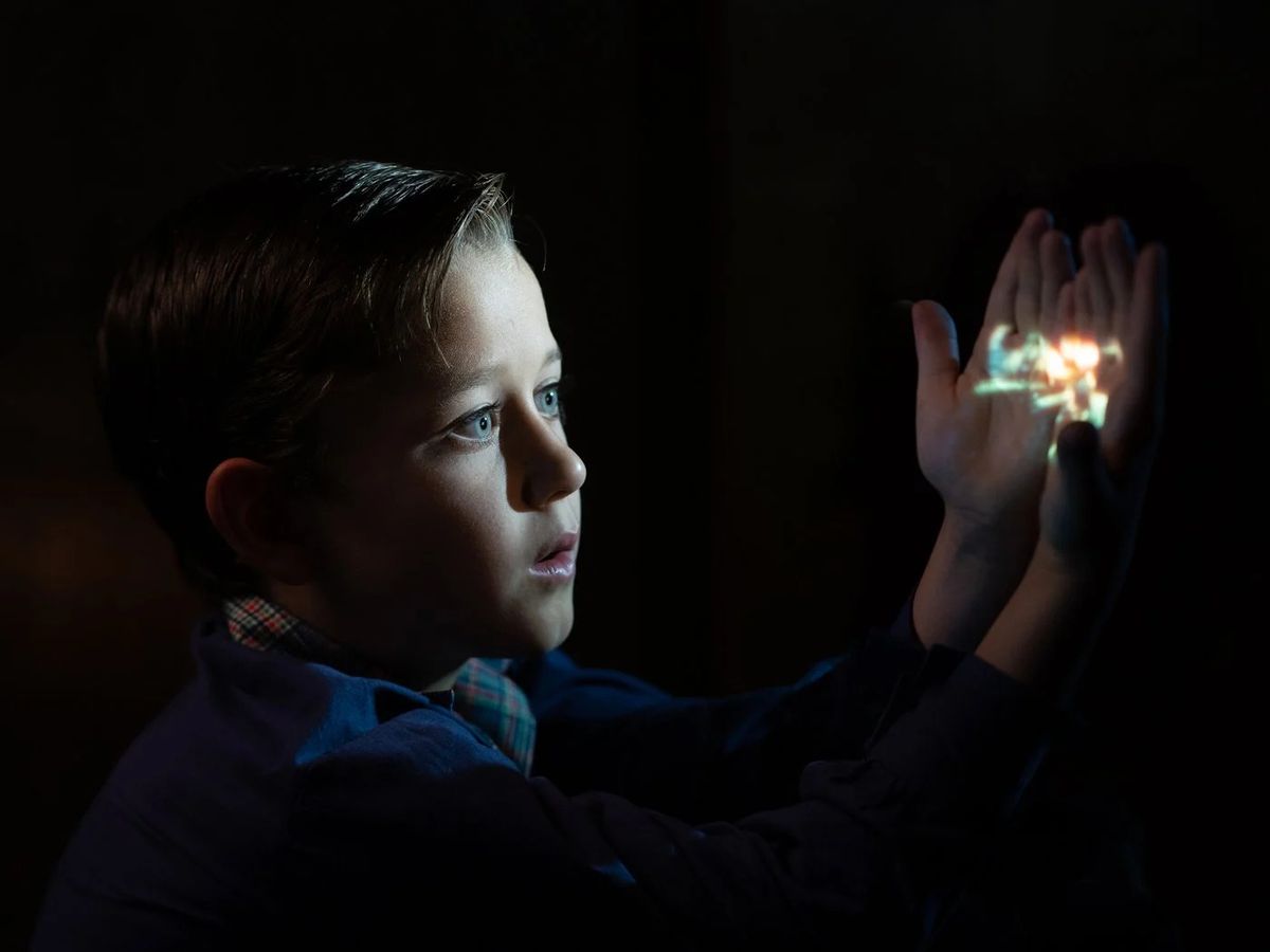 A young boy (Mateo Zoryon Francis-DeFord) holding his hands up in front of the light of a film projector, marveling at an appearance of an image across his fingers.
