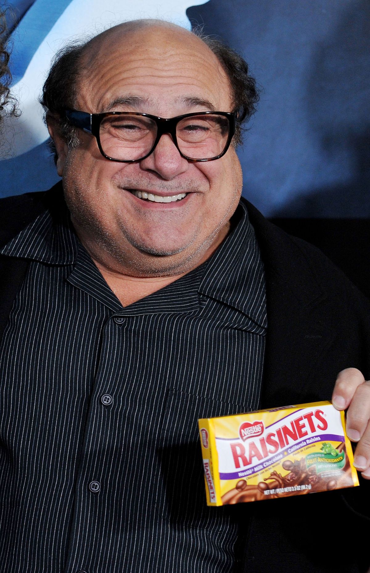 Danny DeVito holds up a box of Raisinets and smiles