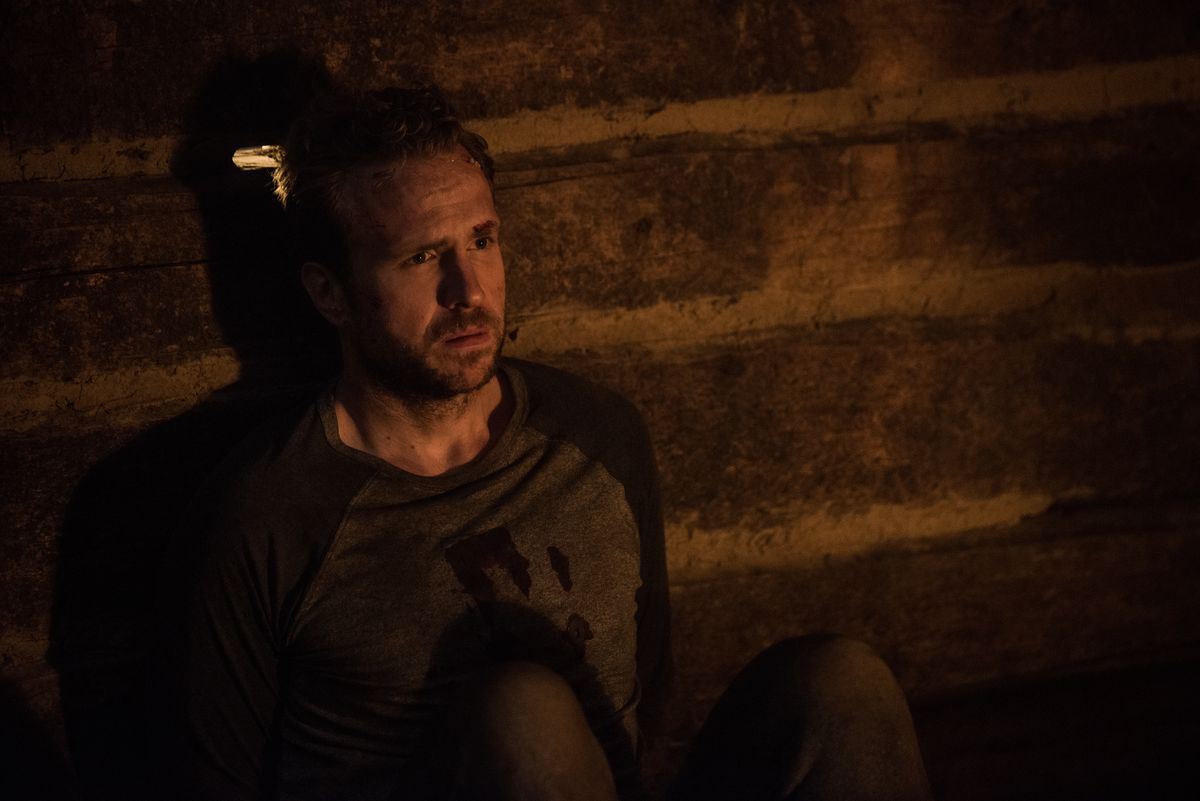 A man wearing a bloodied shirt (Rafe Spall) sits immobilized against a brick wall lit by a nearby fireplace with a look of frustration on his face.