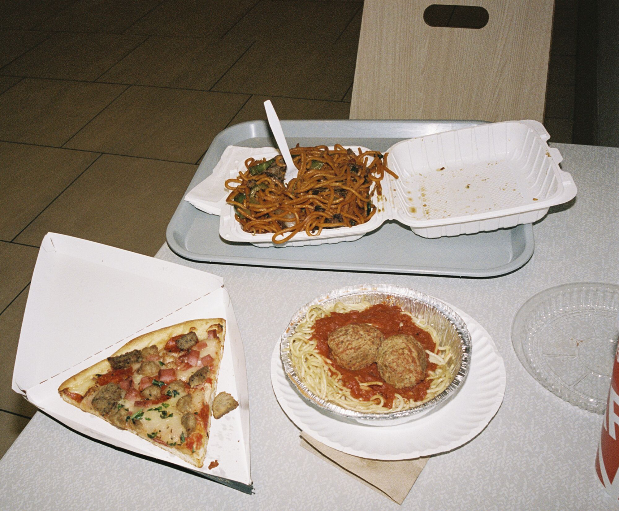 A slice of pizza, an aluminum to-go bowl of spaghetti and meatballs, and a styrofoam container filled with lo mein noodles