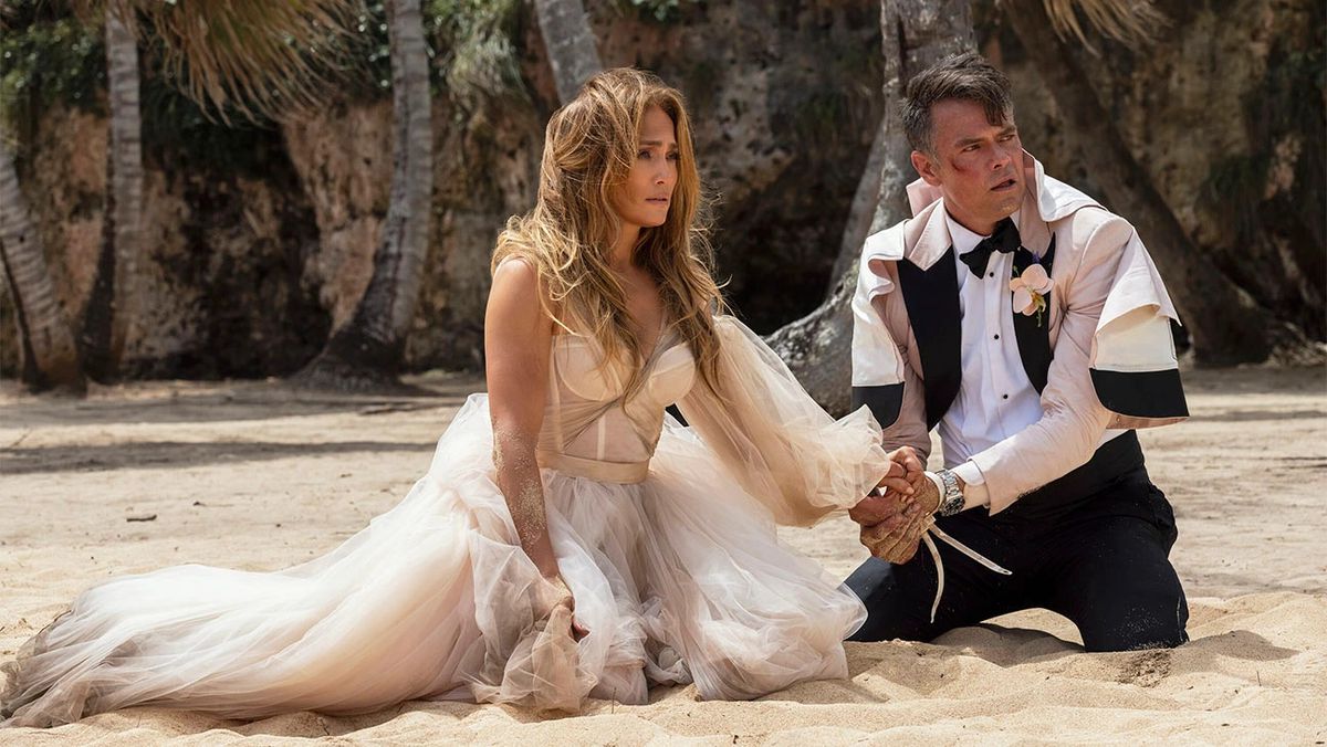 A woman in a torn wedding dress (Jennifer Lopez) kneels on a beach beside a man (Josh Duhamel) in a tattered tuxedo with a bloodied bruise on his cheek.