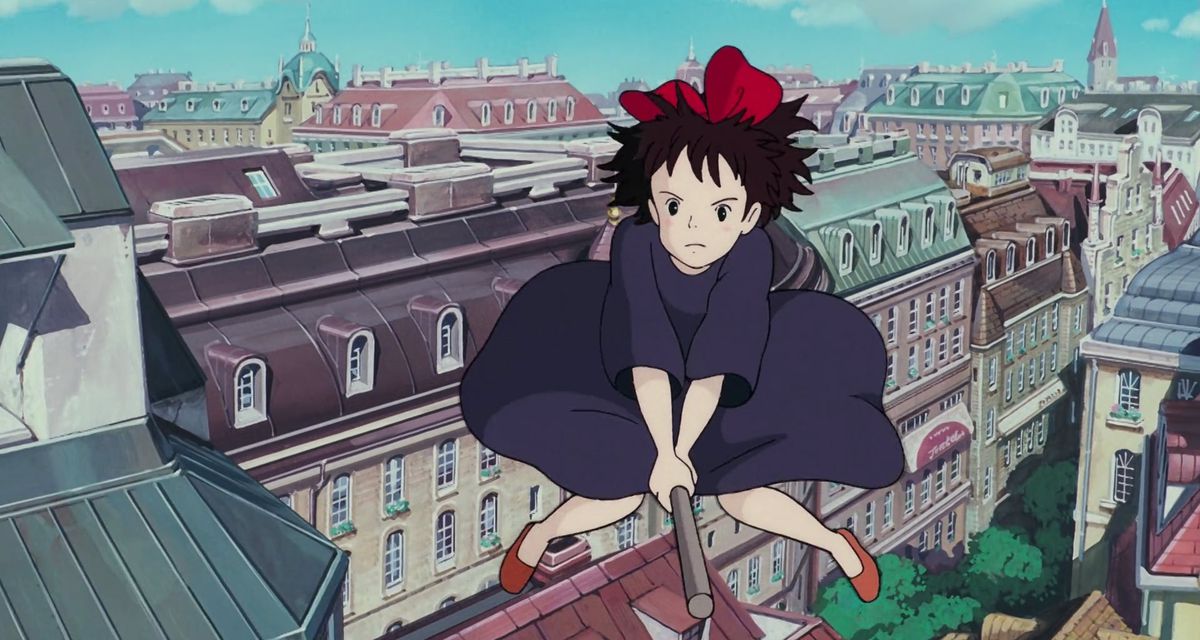 Kiki, an anime girl with black hair and a red bow on top, rides a broom over a European-style cityscape in Studio Ghibli’s Kiki’s Delivery Service.