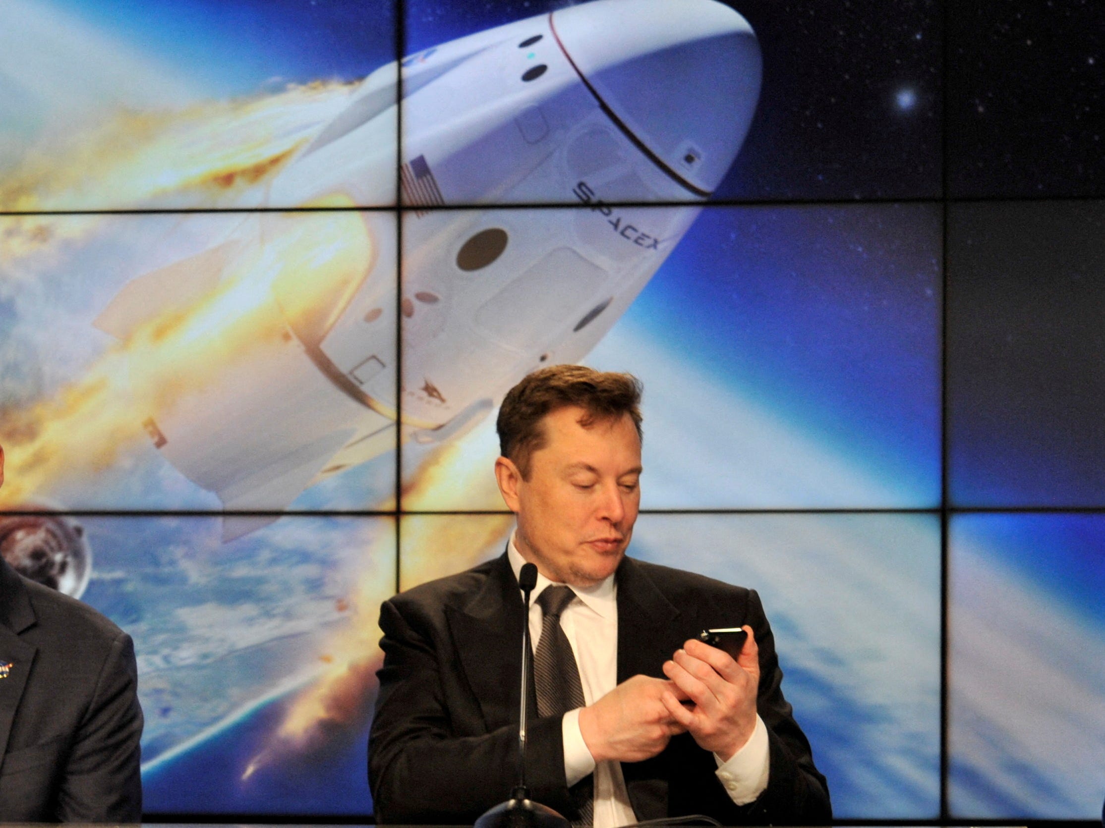 Elon Musk ser på telefonen sin med en skjerm av en romkapsel som går inn i bane bak seg.