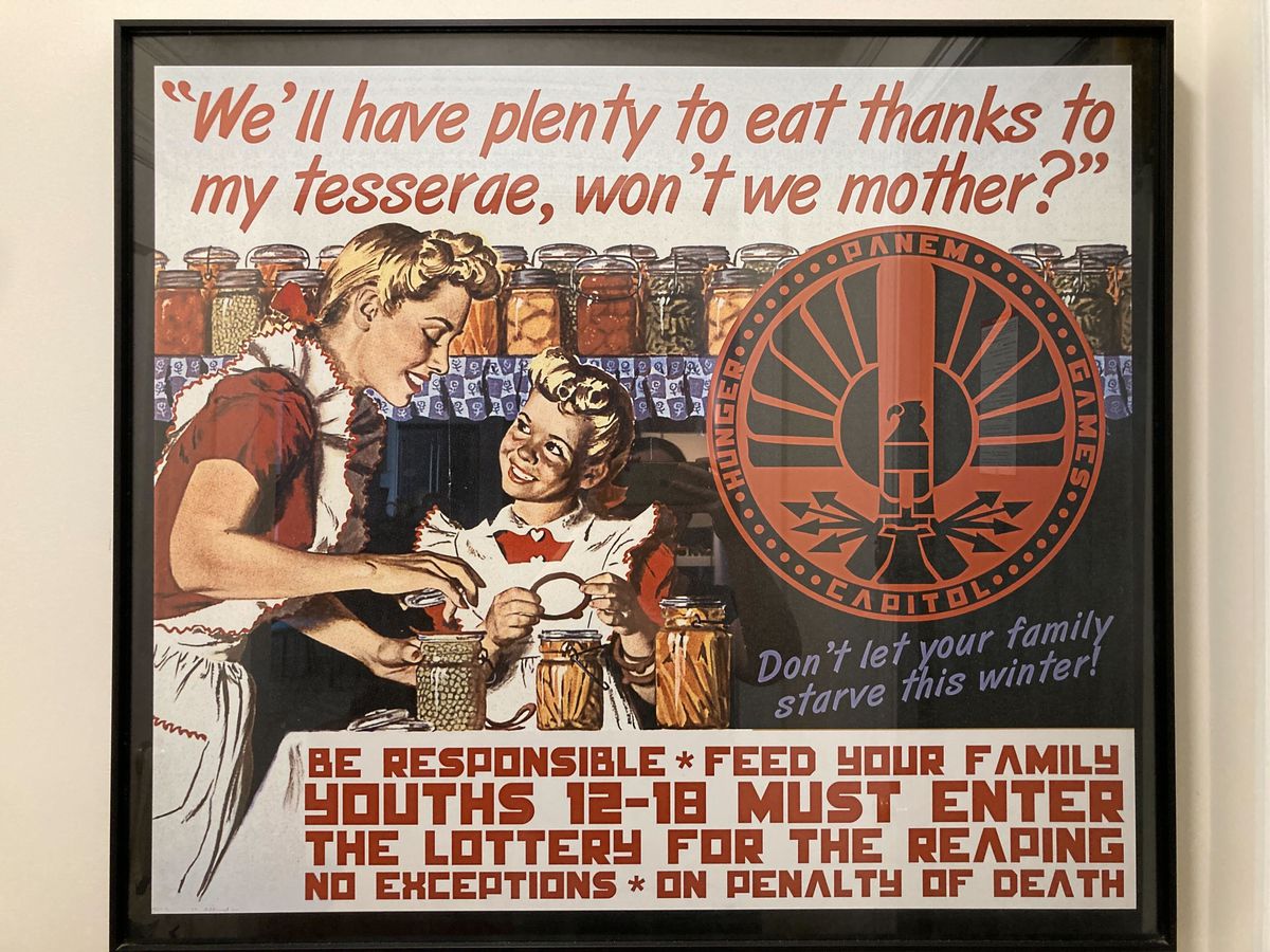A photo of a nearly square framed poster, hung on a wall. A June Cleaver-type cans preserves with her smiling pigtailed daughter, under the text “We’ll have plenty to eat thanks to my tesserae, won’t we mother?” Another bit of text says “Don’t let your family starve this winter!” At the bottom of the poster, block text reads BE RESPONSIBLE — FEED YOUR FAMILY — YOUTHS 12-18 MUST ENTER THE LOTTERY FOR THE REAPING — NO EXCEPTIONS — ON PENALTY OF DEATH.
