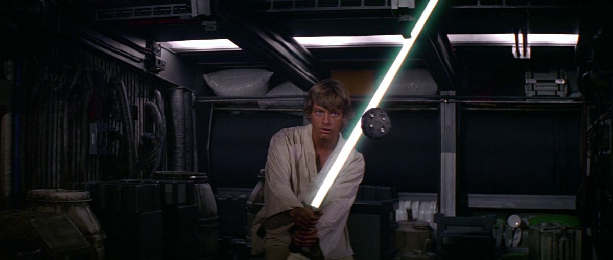 Luke Skywalker drži svoj svetlobni meč nasproti drona za vadbo v filmu Novo upanje.