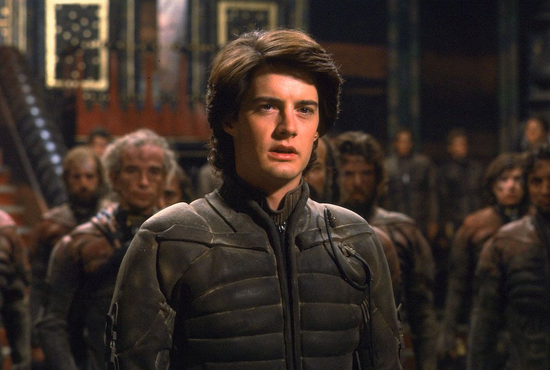 A young man (Kyle Maclachlan) with brown hair dressed in a dusty high-tech suit stands in an atrium surrounded by people dressed in similar suits in Dune (1984).
