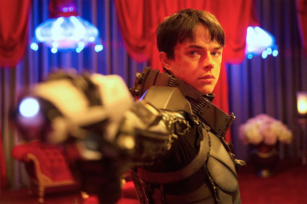 A young man (Dane DeHaan) with short black hair in a high-tech space suit aims a glowing pistol in a red lit room with blue chandeliers in the background in Valerian and the City of a Thousand Planets.