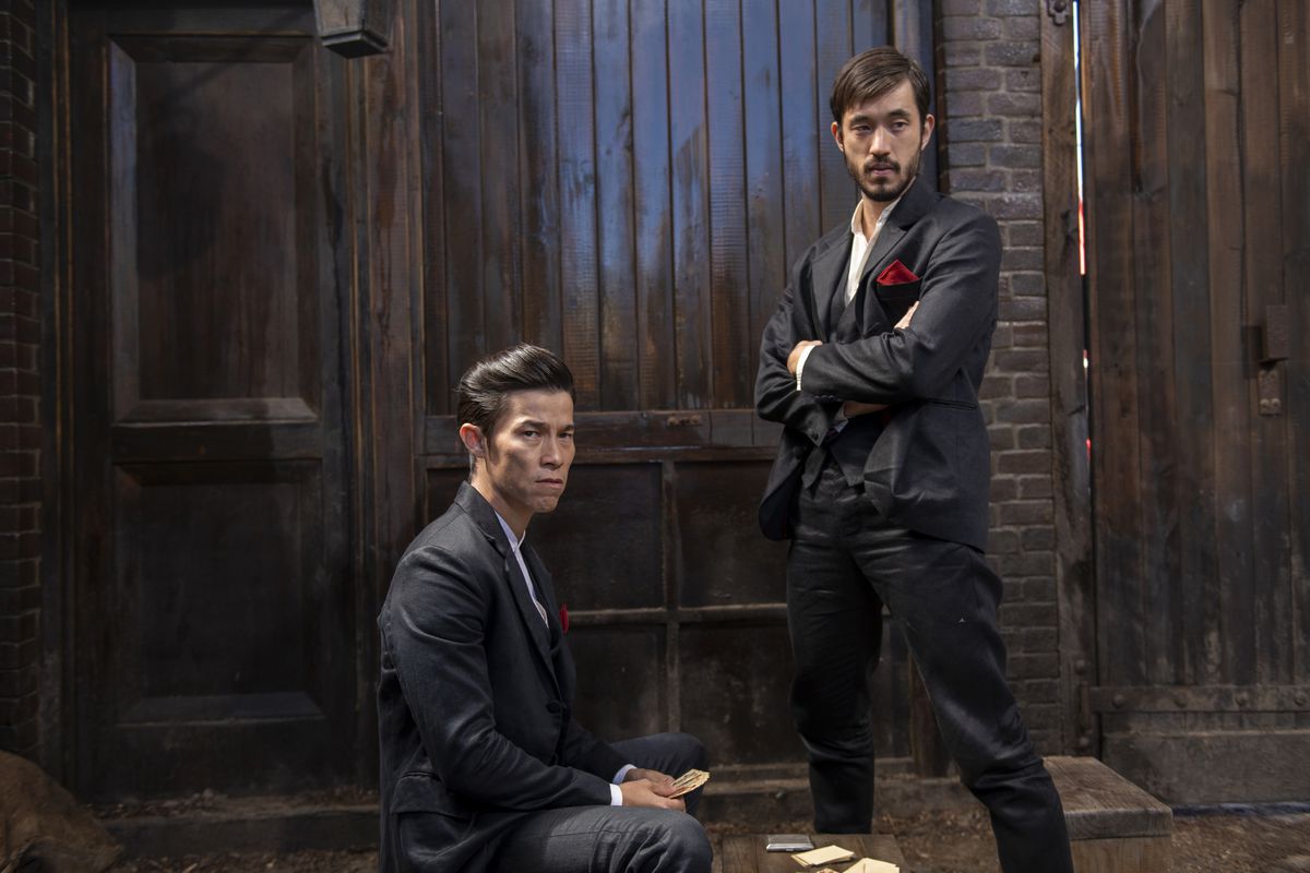 Jason Tobin, sitting, and Andrew Koji, standing with his arms crossed, wear black suits with red handkerchiefs in the pocket in front of a wooden wall in Warrior.
