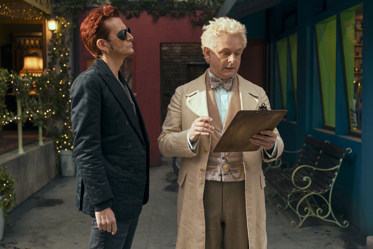 Crowley (David Tennant) stands and looks annoyed at Aziraphale (Michael Sheen) who is looking down at his clipboard