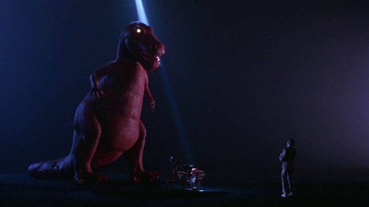 Pee-Wee Herman (Paul Reubens) staring up at a Red T-Rex standing next to a bicycle surrounded by darkness in Pee-Wee’s Big Adventure.