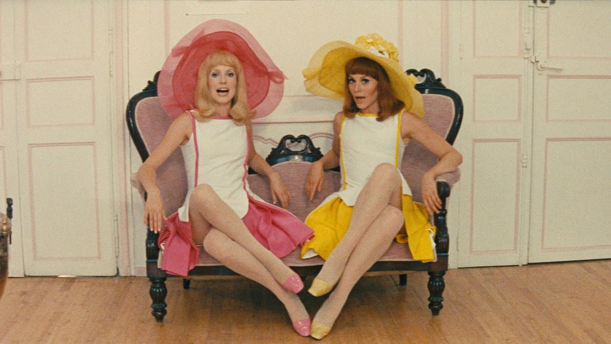 (L-R) Delphine (Catherine Deneuve) and her sister Solange (Françoise Dorléac) seated on a couch in pink and yellow outfits in The Young Girls of Rochefort.