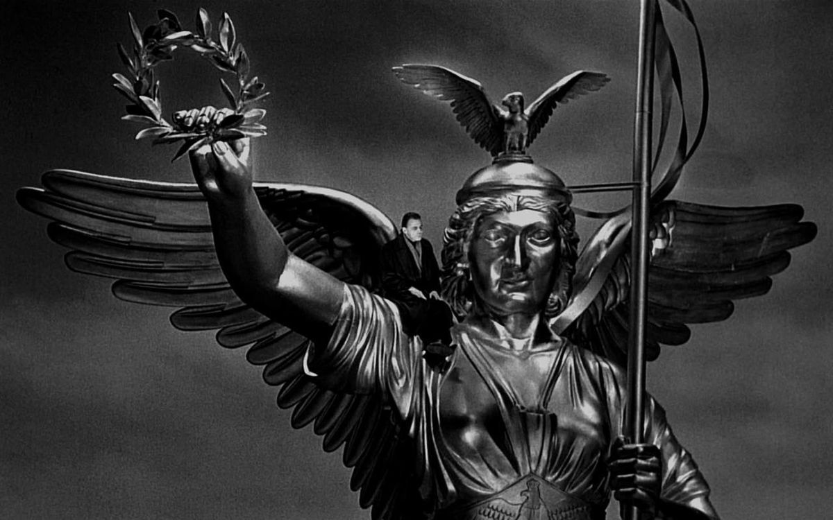Bruno Ganz as an angel sitting on the shoulder of a winged statue in a helmet holding a spear and a wreath in Wings of Desire.