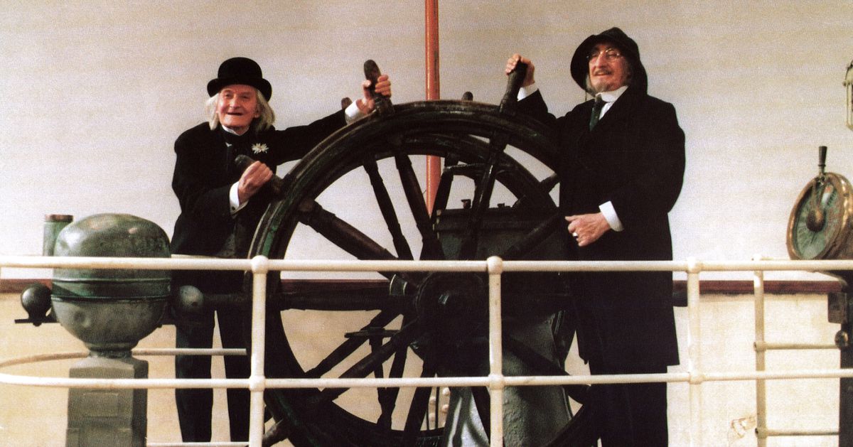 Two elderly men standing with their hands on the wheel of a ship in And The Ship Sails On.