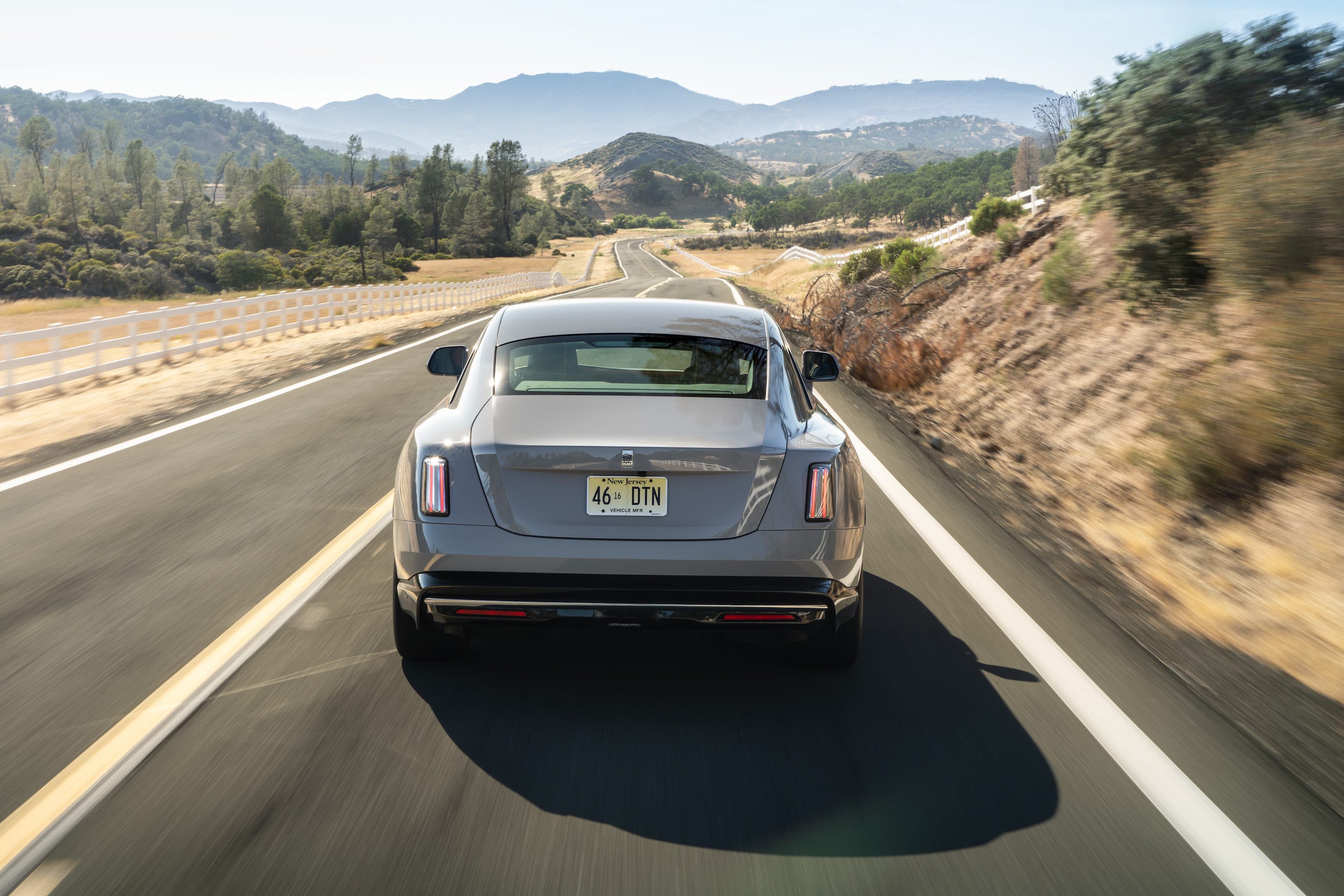 A gray Rolls-Royce Spectre electric car viewed from the rear, driving down a road with mountains in the distance.