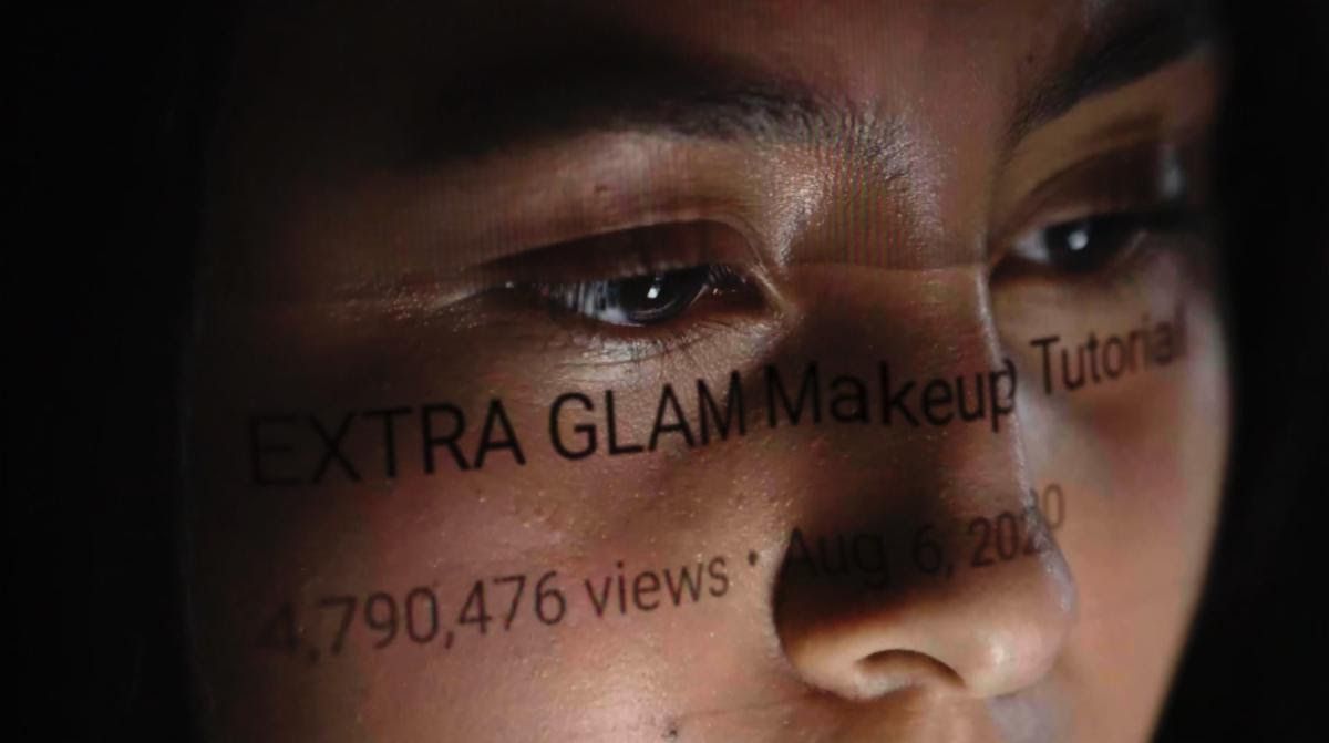 A young person’s face as the words “EXTRA GLAM MAKEUP Tutorial; 4,790,476 views; 2020” play across their face like a reflection of the screen.