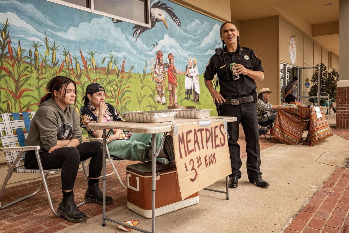 Devery Jacobs, Paulina Alexis, and Zahn McClarnon gather around a table that advertises Meatpies for $3 each, in front of a mural, in Reservation Dogs.