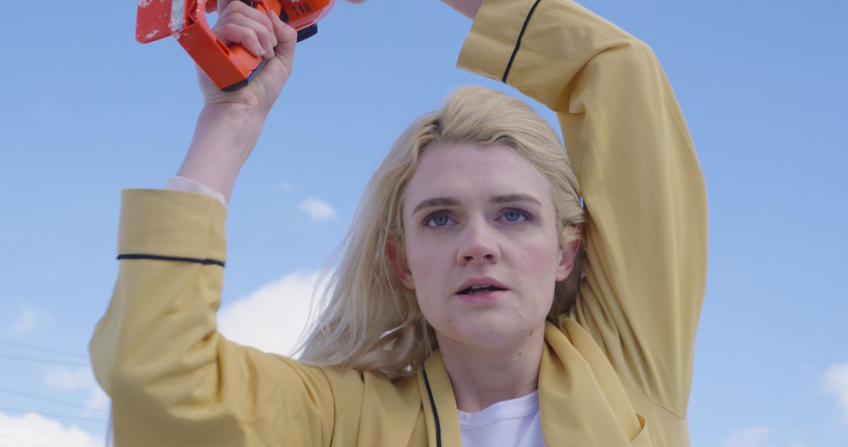 Gayle Rankin in a yellow coat holding a chainsaw with an orange handle over her head in Bad Things.