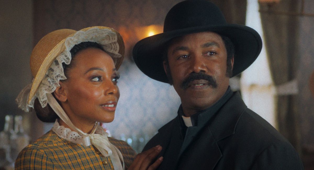 Michael Jai White, wearing a preacher’s collar and a black cowboy hat, stands next to Erica Ash, wearing a bonnet under a sun hat, in Outlaw Johnny Black.