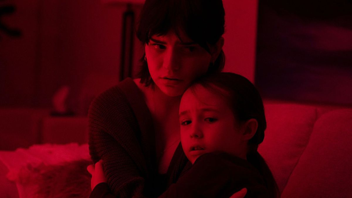 (L-R) Sophie Thatcher and Vivien Lyra Blair huddle close together on a couch in the dark bathed in a deep red light in The Boogeyman.