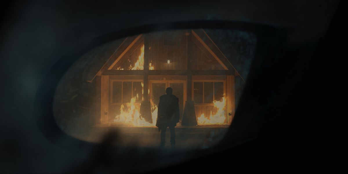 The image of a person standing in front of a burning building reflected in a rearview mirror.