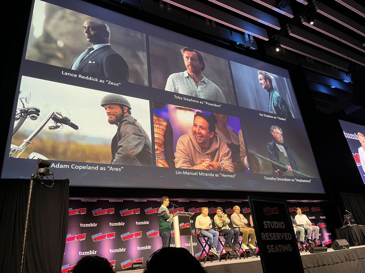A photo of the stage at NYCC with the Percy Jackson crew sitting in chairs in front of a large screen that has photos of the adult actors and their titles: “Lance Reddick as Zeus”, “Toby Stephans as Poseidon”, “Jay Duplass as Hades”, “Adam Copeland as Ares”, “Lin-Manuel Miranda as Hermes”, and “Timothy Omundson as Hephaestus”