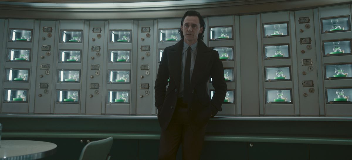 Loki (Tom Hiddleston) stands against a bunch of screens with his hands in his pockets