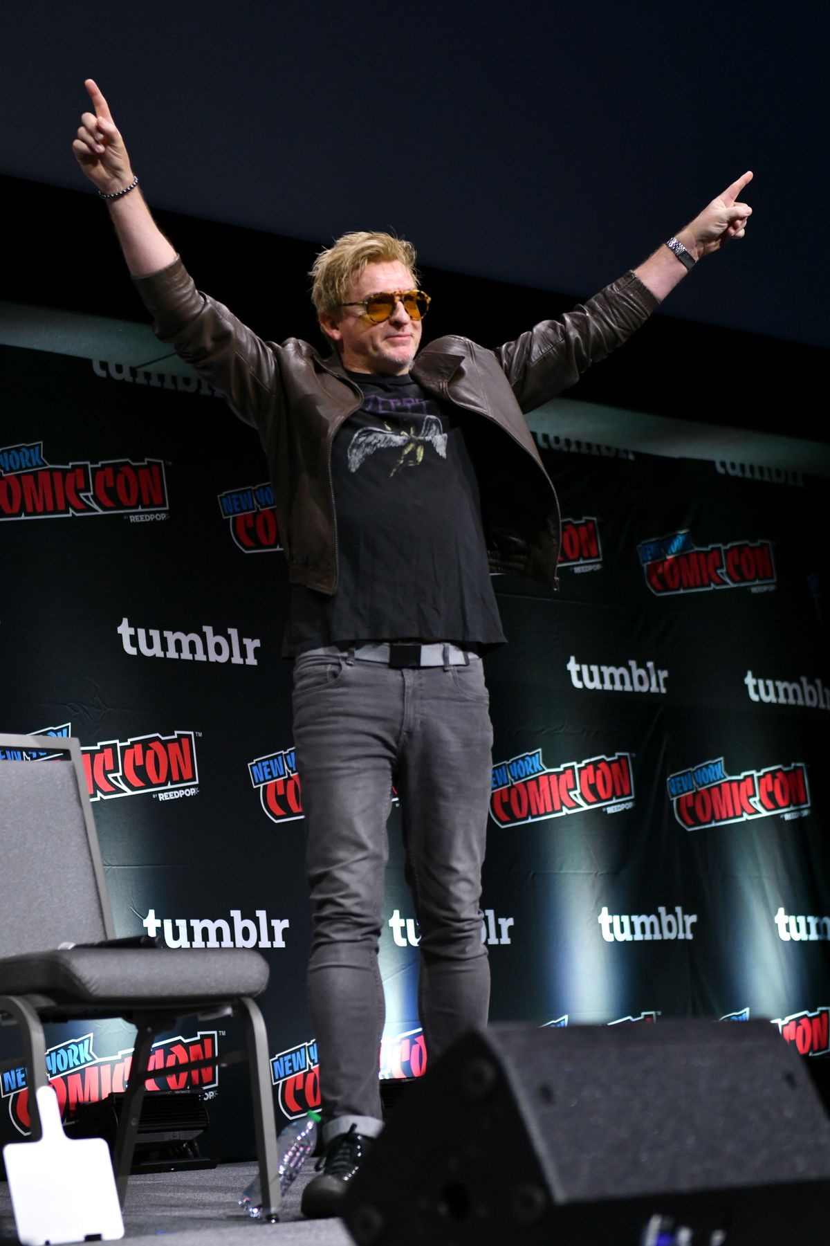 Rhys Darby takes the NYCC 2023 stage in rock star glasses holding his hands up and pointing to the audience