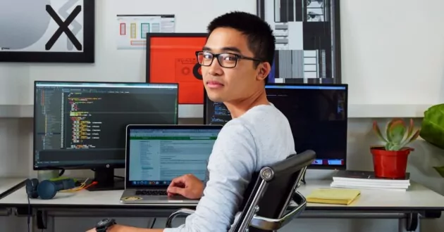 Молода людина в окулярах сидить у робочому кріслі перед трьома екранами комп’ютера, стикається з камерою та посміхається
