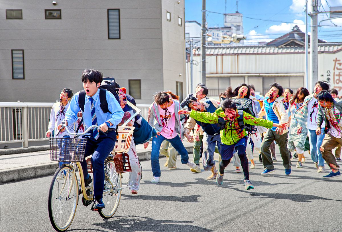Eiji Akaso näyttelee Akiraa ajamassa polkupyörällä pois zombilaumasta Zom 100: Bucket List of the Dead -pelissä.