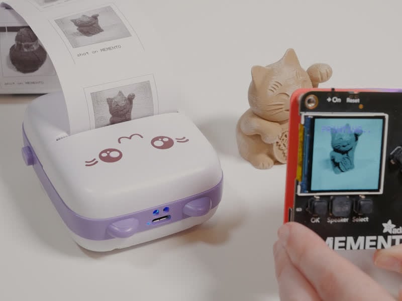 a memento taking a picture of a 3d printed lucky cat model that is printing on a cat thermal printer