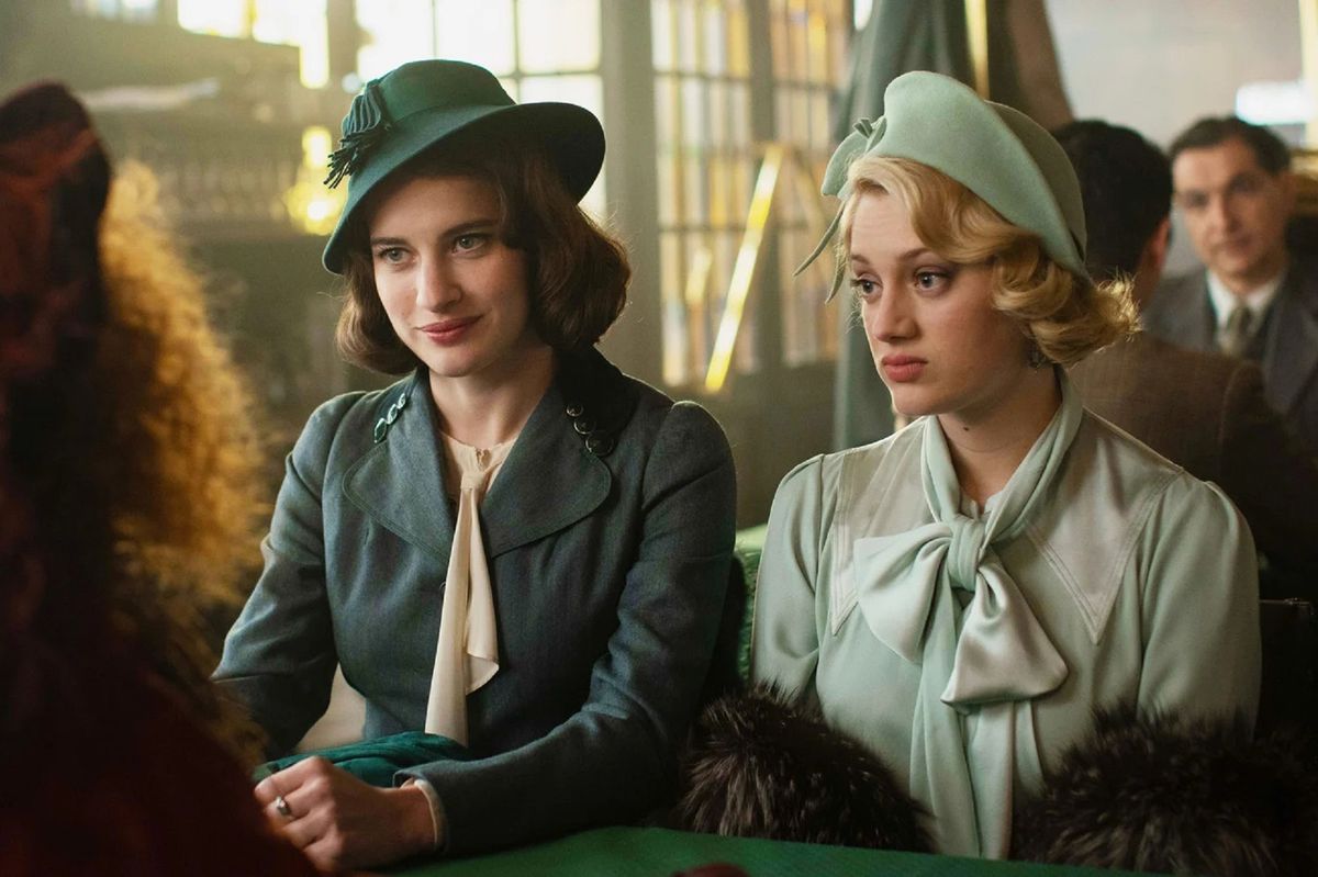Two women in green coats and hats sitting next to one another at a table.