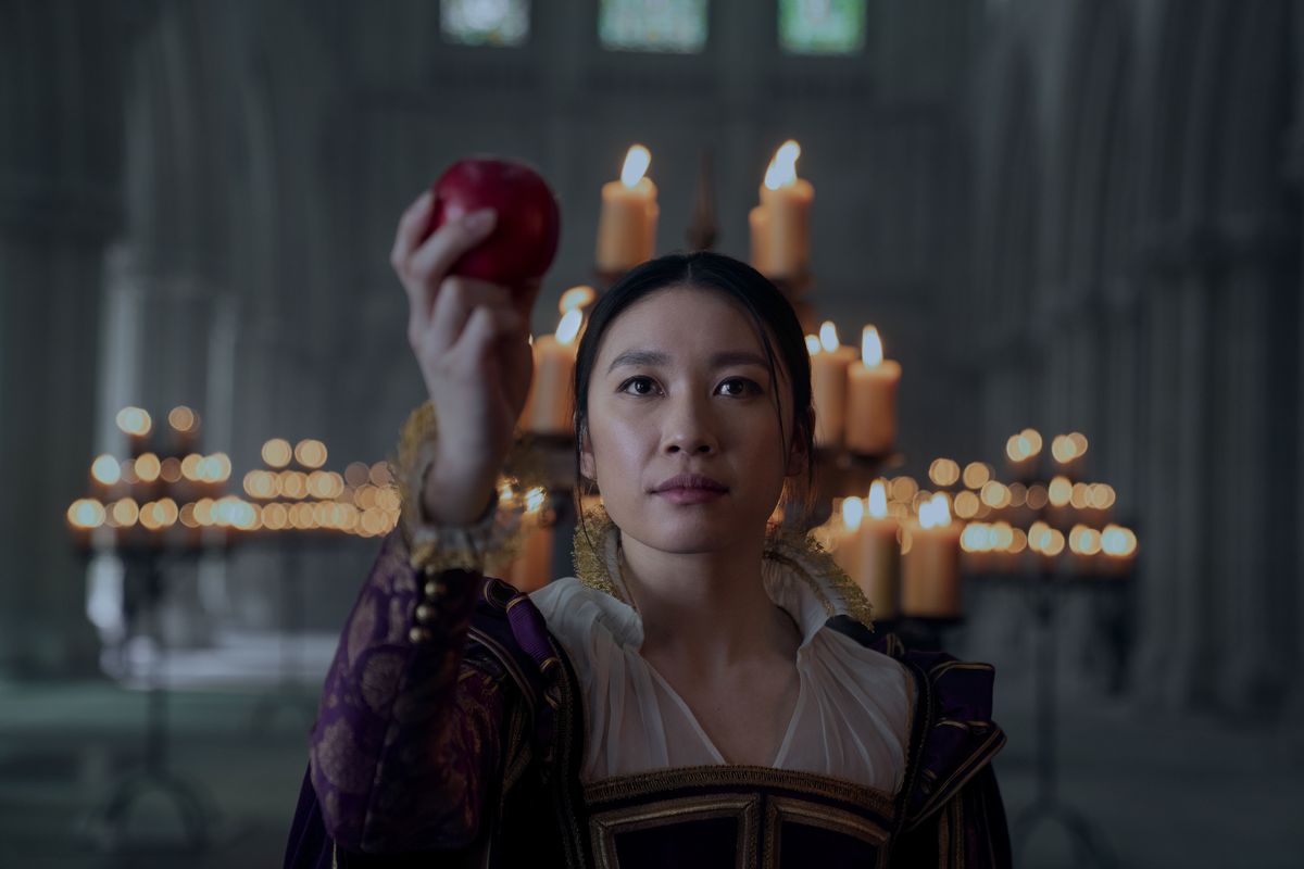 Jess Hong as Jin wearing Victorian era clothing and holding up an apple in a throne room