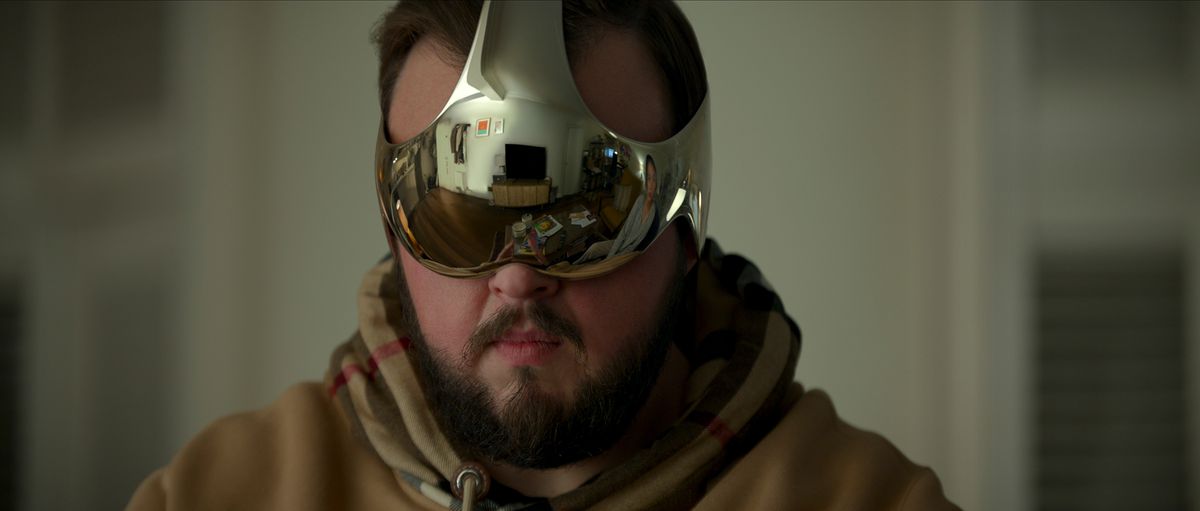 A bearded man wears a highly reflective futuristic VR device that reflects the image of the character Jin in 3 Body Problem