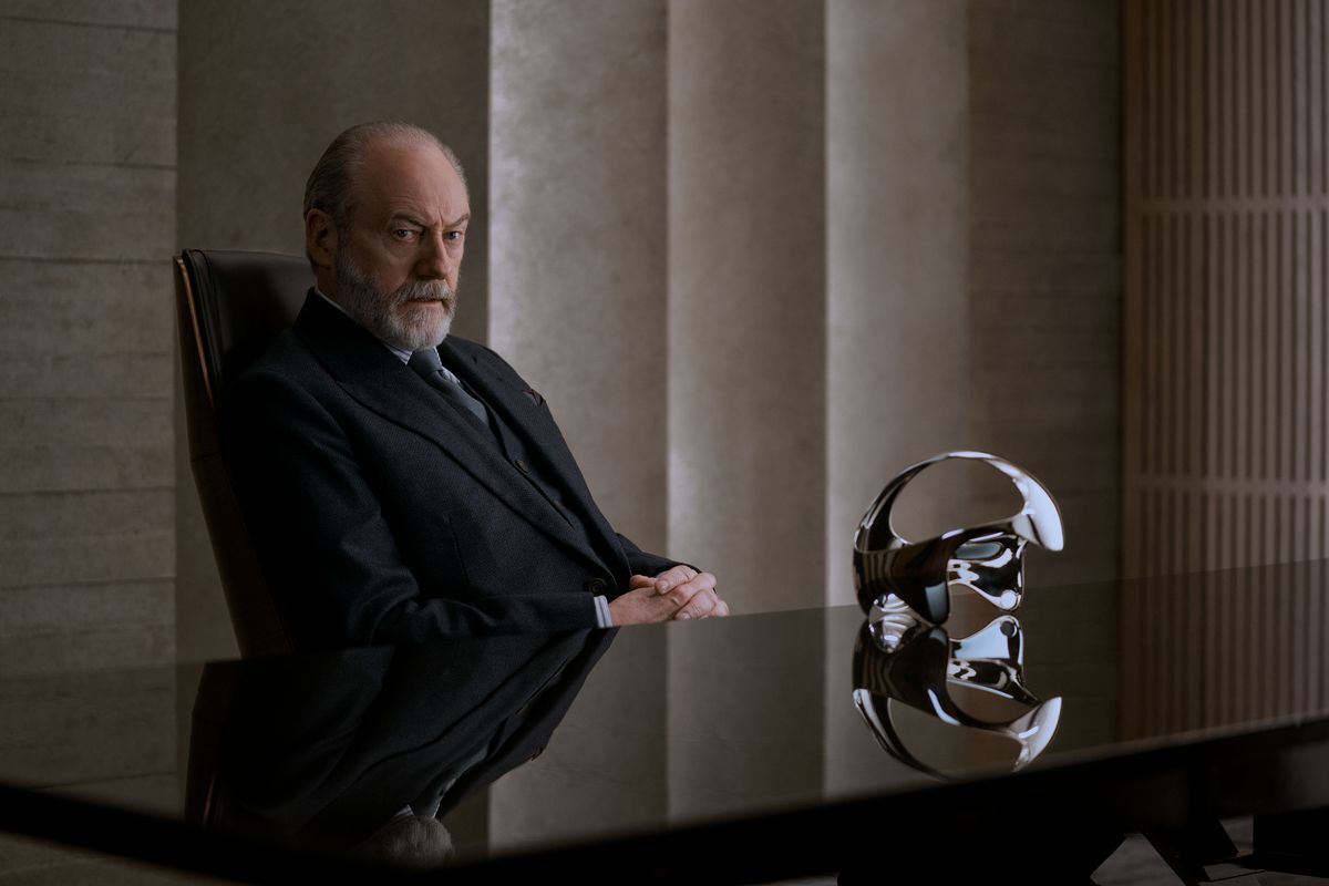 Thomas Wade (Liam Cunningham) sitting at a desk with a futuristic headset on a table.