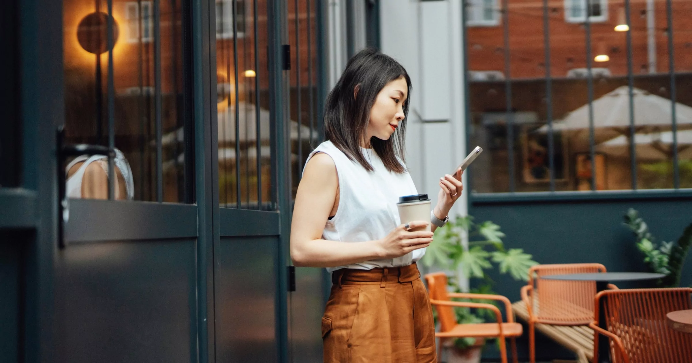 Person standing outside cafe with coffee in one hand and cell phone in the other, looking at cell