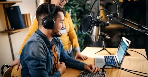 Two coworkers smiling at computer, one sitting in chair and wearing headphone, the other standing