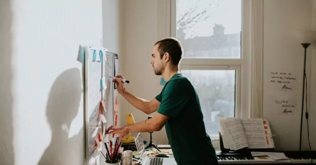 Mężczyzna pochyla się nad zabałaganionym biurkiem i pisze na tablicy zamontowanej na ścianie w domowym biurze