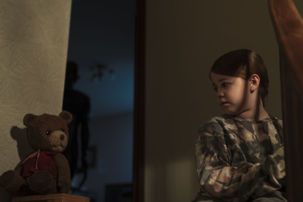 A young girl played by Pyper Braun sits at the top of the stairs next to a teddy bear while an ominous shadowy figure lurk behind her in Imaginary