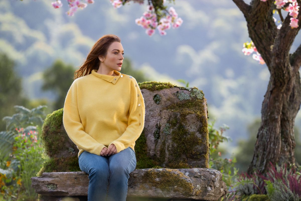 Lindsay Lohan in Irish Wish sitting on a stone bench under a pretty tree. She wears a yellow sweater, red hair blowing in the wind, as she looks to the right.