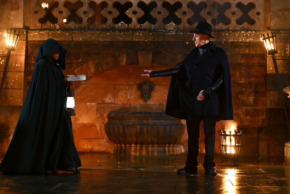 Alan Cumming accepting an envelope from a hooded traitor on The Traitors season 2