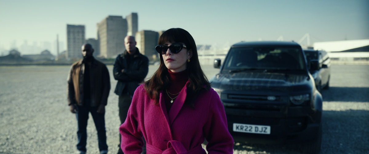 Kaya Scodelario as Susie Glass stands in front of a car in The Gentleman wearing a pink coat