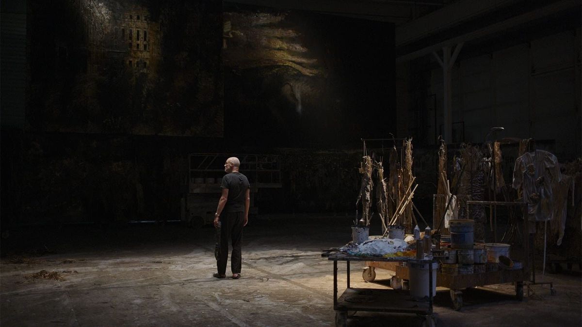 Een man die in een verduisterde studio staat, omringd door kunstbenodigdheden en grote schilderijen.