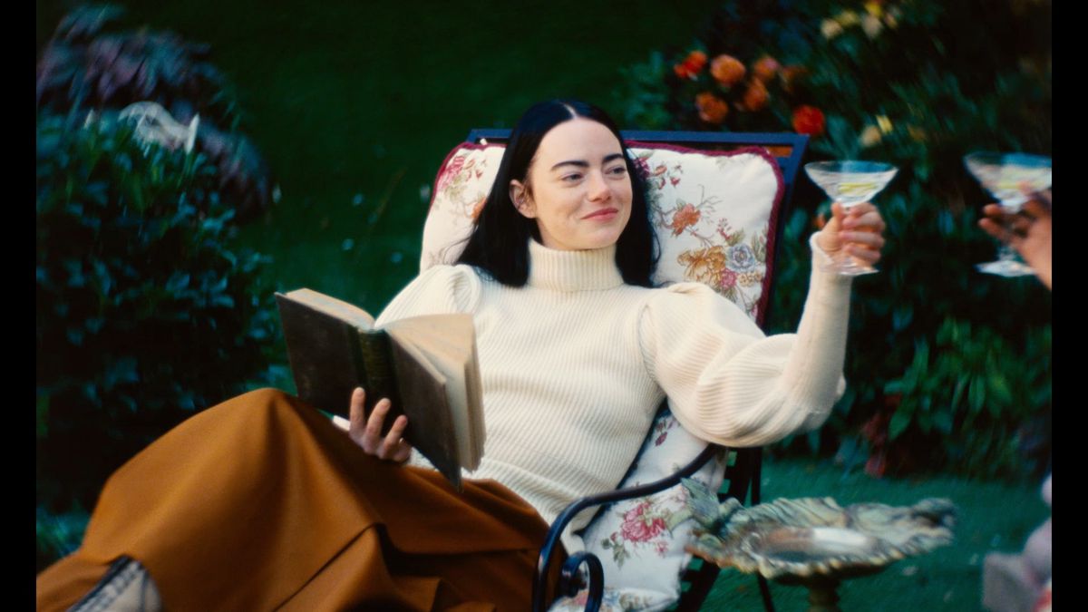 Emma Stone como Bella Baxter descansando em uma cadeira de jardim, segurando um livro, sorrindo e levantando uma taça de coquetel em Poor Things
