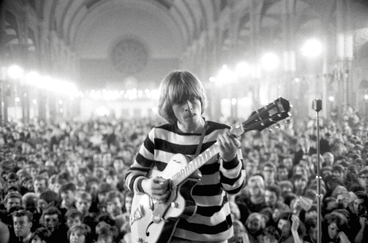 Foto de Brian Jones em frente a um teatro lotado tocando guitarra em The Stones & Brian Jones.