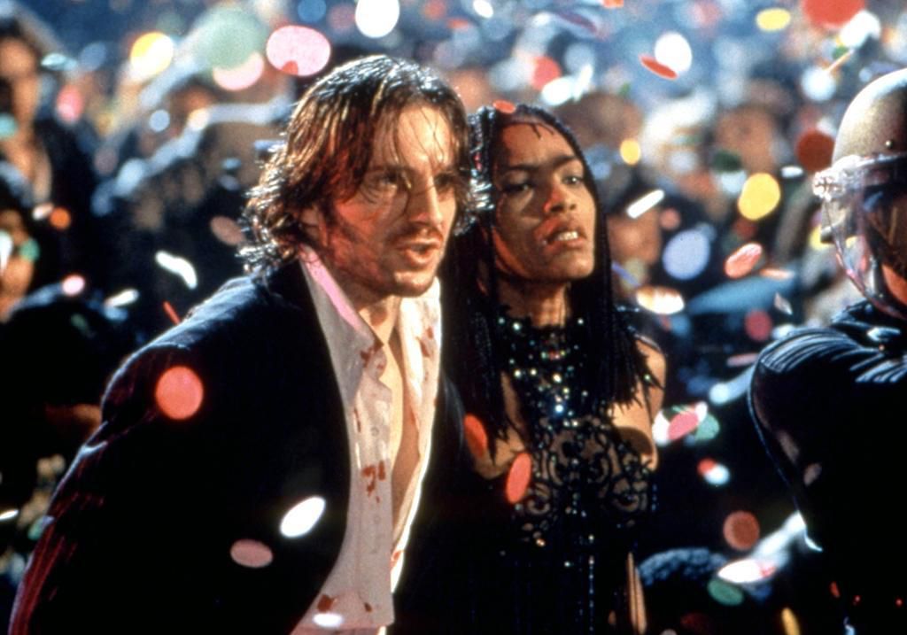 A disheveled man and a woman in a black dress stand in a large crowd while confetti rains down from the sky.