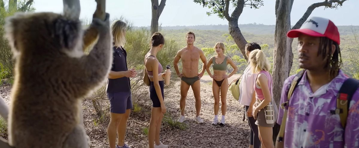 A group on a hike in Australia. In the forefront, a Black man stars, transfixed, at a koala. Meanwhile a hunky blonde man wearing only briefs and a beautiful blonde woman stand with their arms akimbo in the background. Everyone else in the group gawks at them.