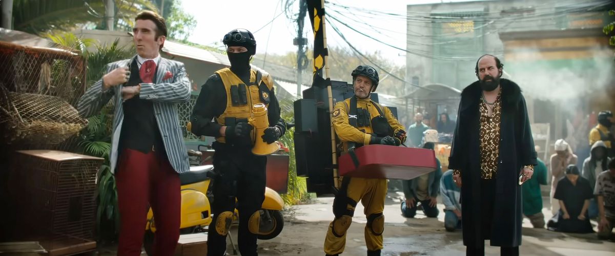 Some of the baddies in Boy Kills World stand in a run-down slum they’re terrorizing, with yellow-and-black-clad soldiers holding a crowd of locals down on their knees in the background. In the foreground: a couple more of those soldiers, nattily dressed dandy Glen (Sharlto Copley, in red pants, black vest, and blue striped suit jacket), and thuggish Gideon (Brett Gelman, in a big black open-fronted fur coat).